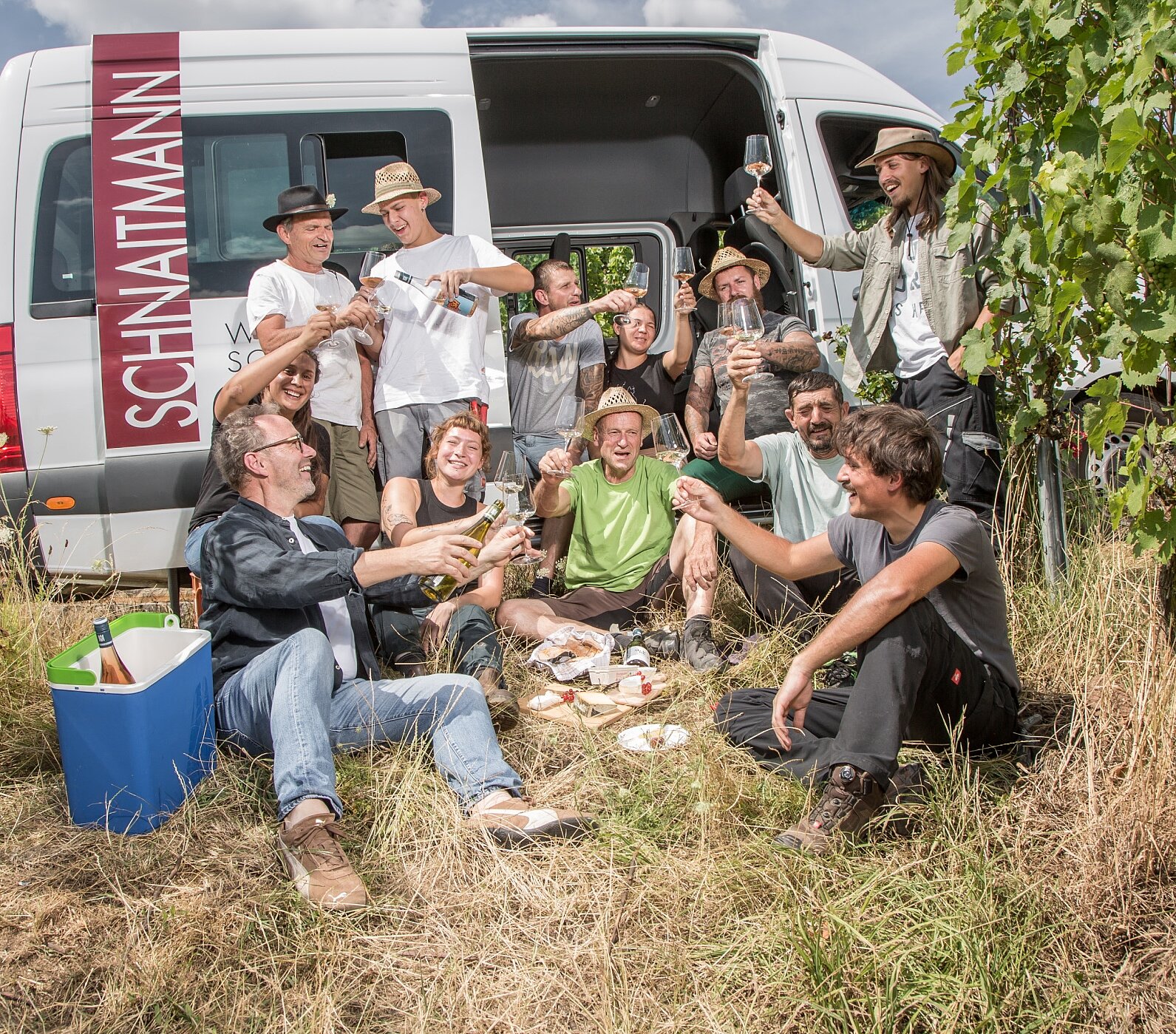 Eine Gruppe von Menschen sitzt und steht in einem Weinberg, lächelt und feiert mit Getränken und Essen vor einem Lieferwagen mit der Aufschrift "Schnaitmann". Es scheint, als würden sie an einem sonnigen Tag ein Picknick zwischen Weinreben genießen.