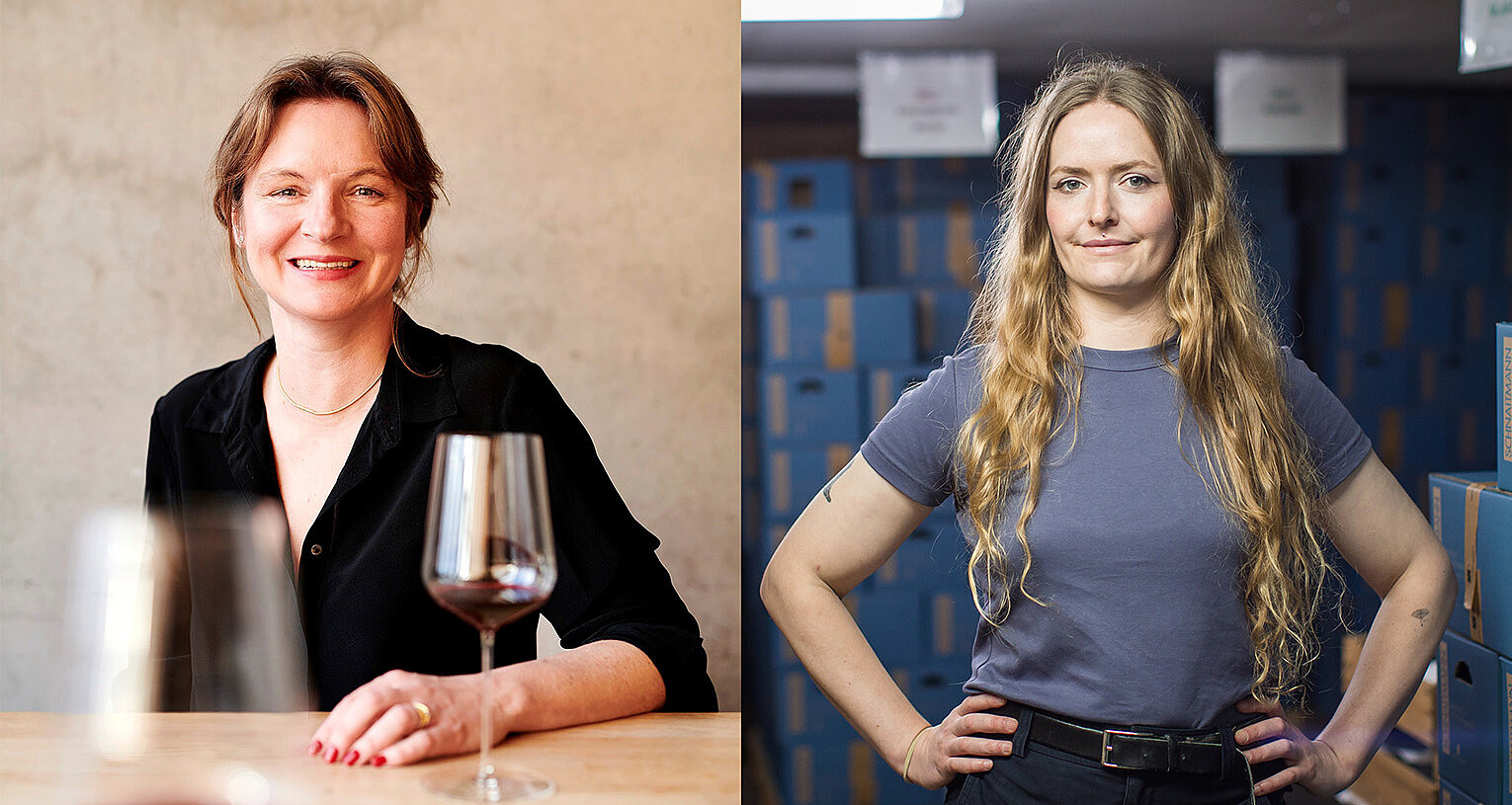 Eine Frau sitzt links im Bild vor zwei Weingläser und eine Frau steht rechts im Bild in einem Weinlager. 