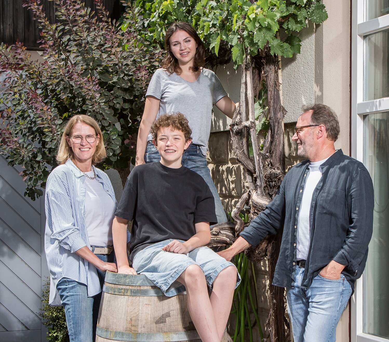 Vier Personen, zwei Erwachsene und zwei Kinder, posieren an einem sonnigen Tag lächelnd im Freien neben einem Holzfass und einer Weinrebe. Ein Kind sitzt auf dem Fass, während die anderen dicht beieinander stehen und glücklich und entspannt aussehen.