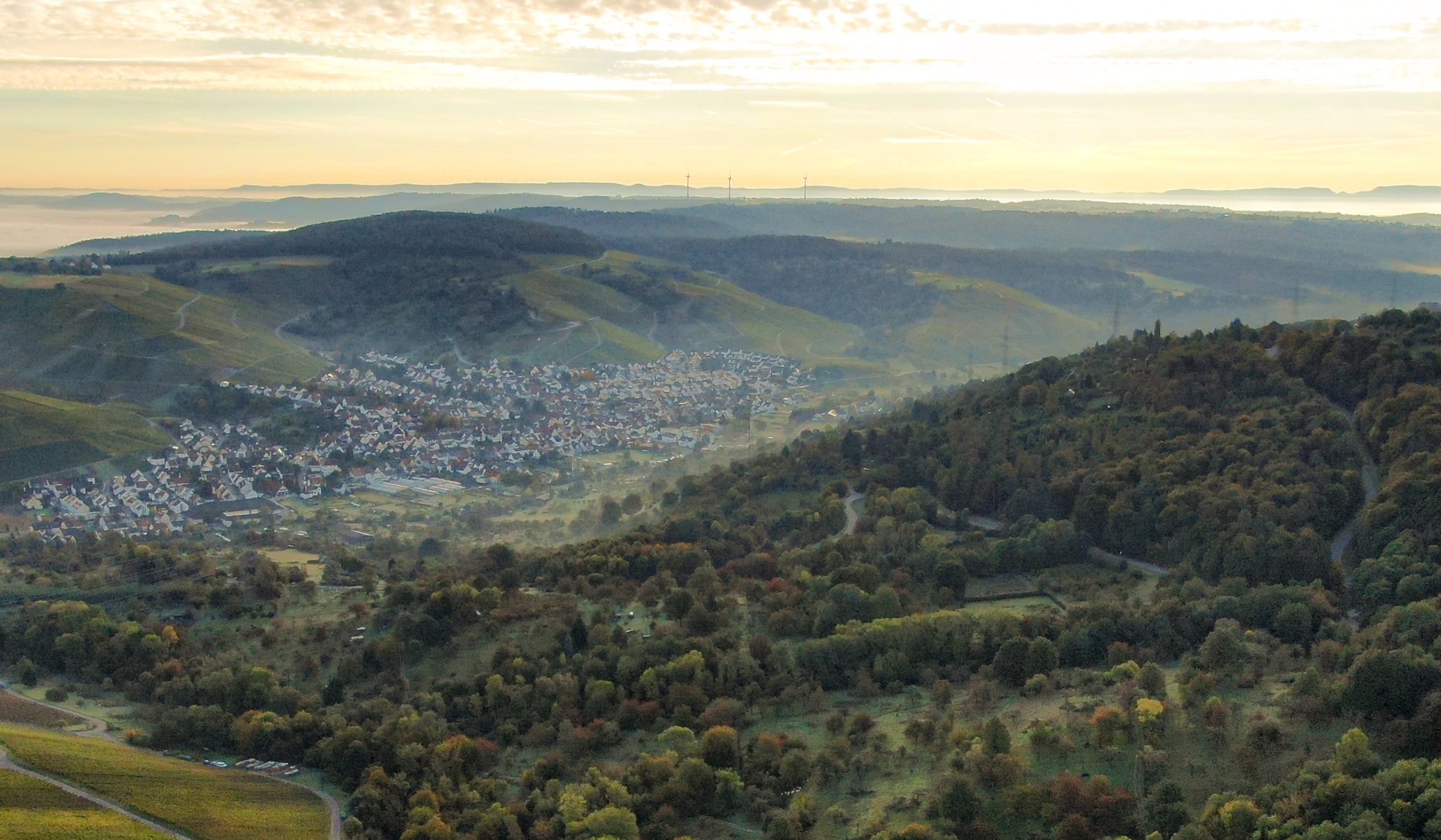 Luftaufnahme einer kleinen Stadt inmitten grüner, sanfter Hügel und Wälder bei teilweise bewölktem Himmel bei Sonnenaufgang oder Sonnenuntergang. Die Landschaft besteht aus einer Mischung aus offenen Feldern, bewaldeten Gebieten und verstreuten Häusern.