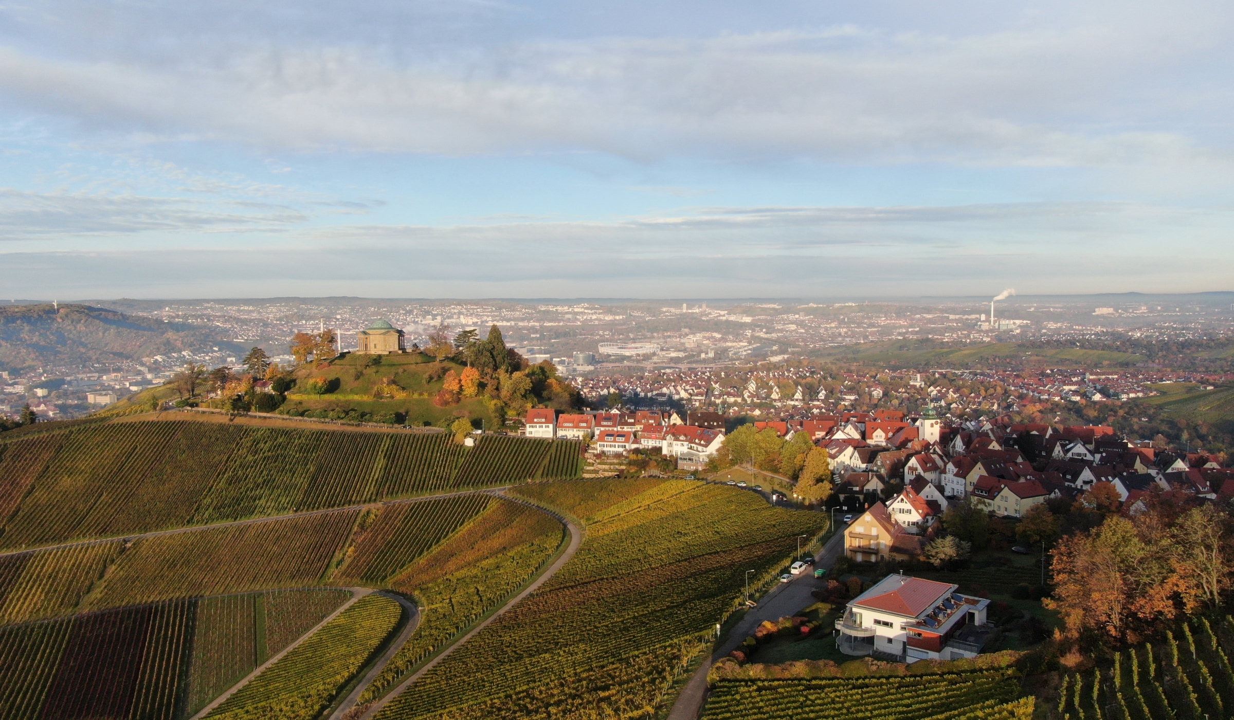Luftaufnahme von sanft geschwungenen Weinbergen und Häusergruppen an einem Hang, mit einem schlossähnlichen Gebäude und einer Stadtlandschaft in der Ferne unter einem teilweise bewölkten Himmel.