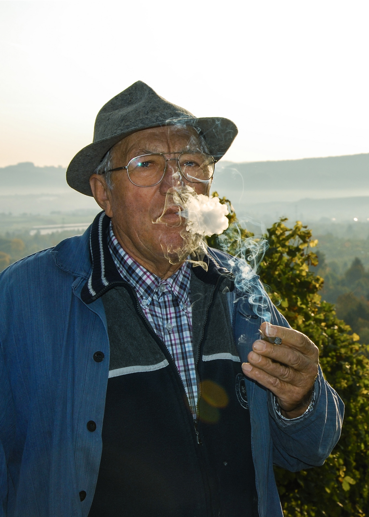 Ein älterer Mann, der eine Brille, einen Hut und eine blaue Jacke trägt, steht im Freien und atmet den Rauch einer Cigarillo aus. Im Hintergrund sind Bäume und eine neblige Landschaft zu sehen.