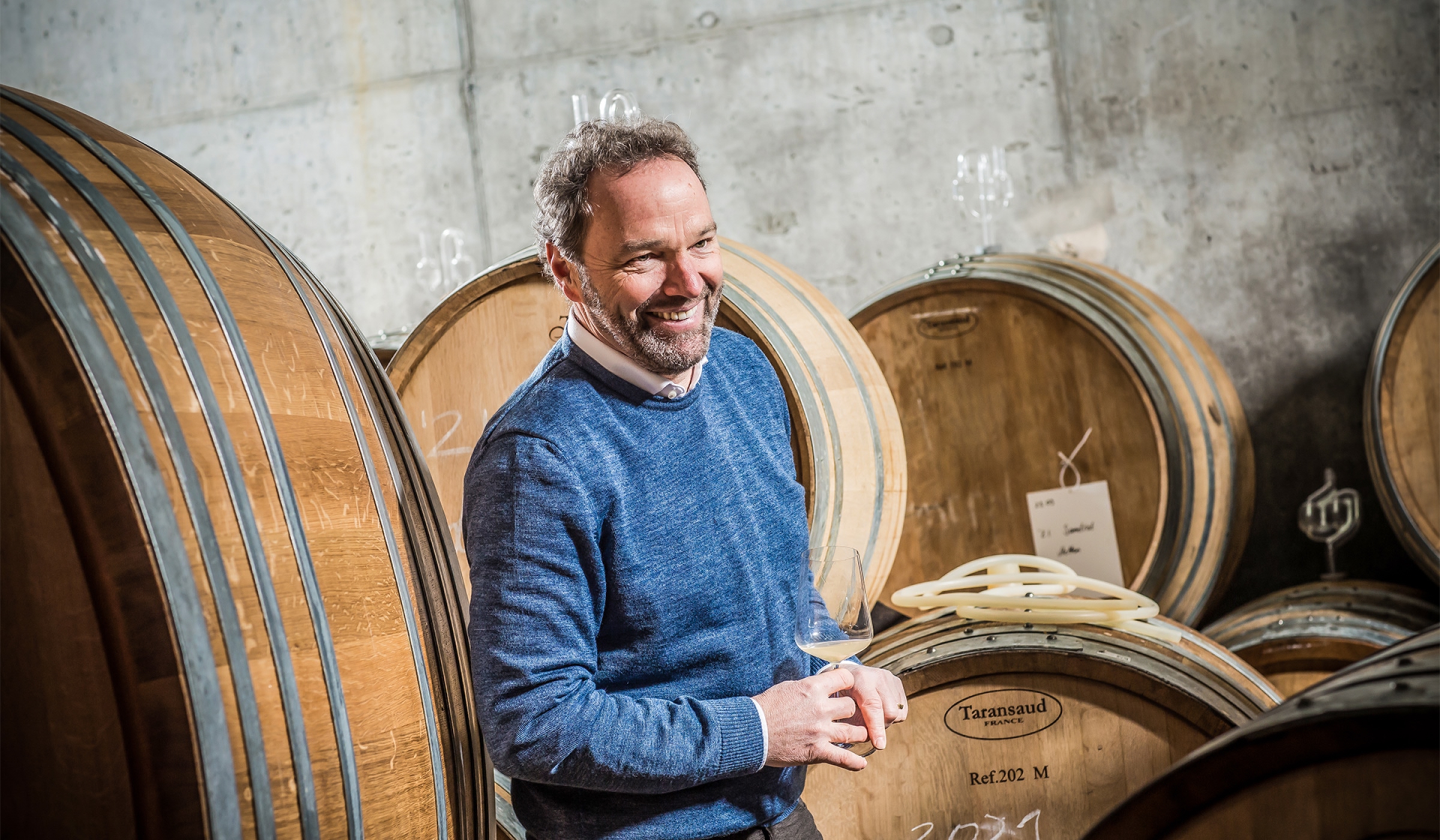 Ein lächelnder Mann in einem blauen Pullover hält ein Weinglas in der Hand, während er zwischen großen Holzfässern in einem Weingut oder Keller steht.