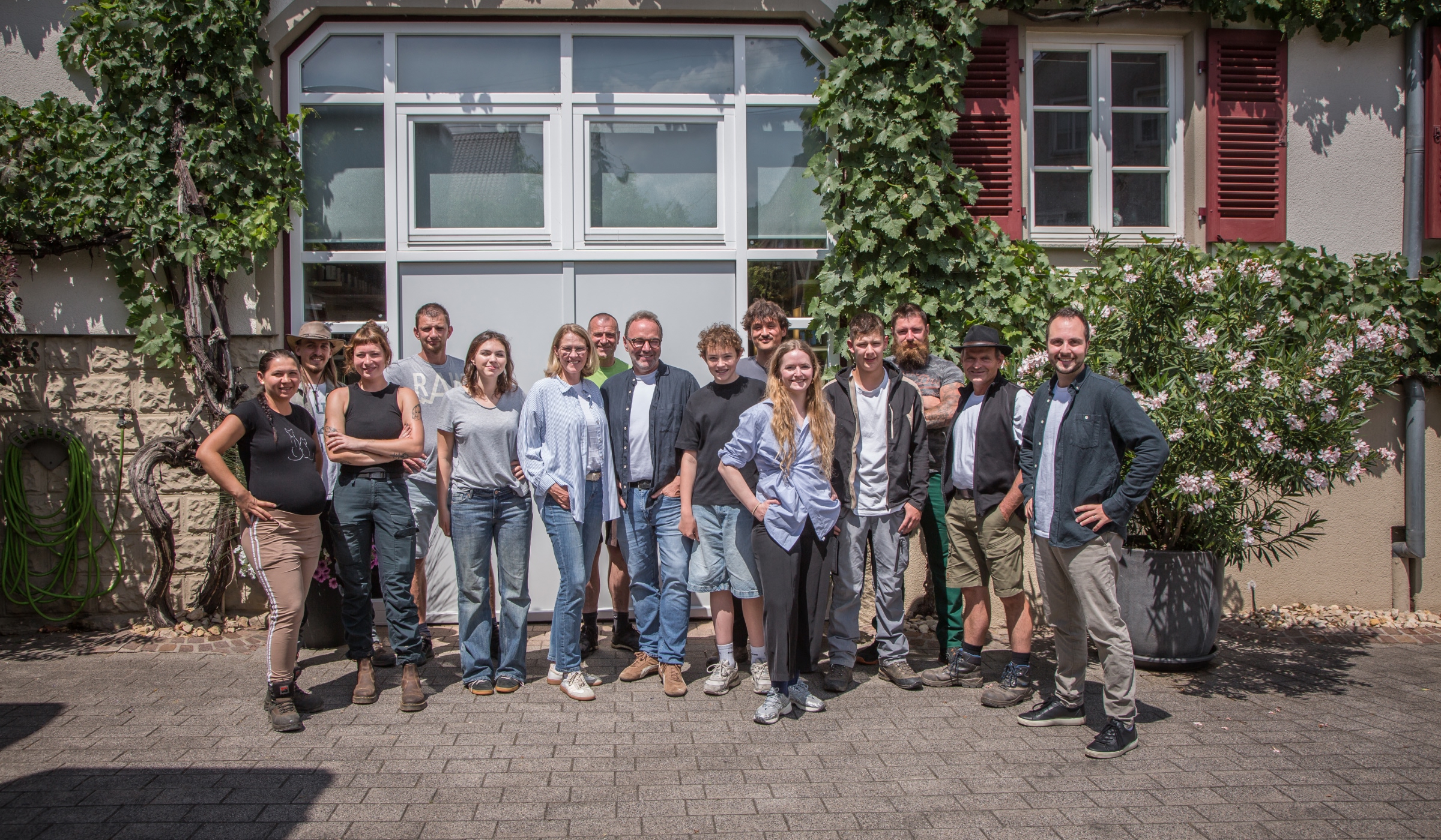 Eine Gruppe von fünfzehn Personen steht an einem sonnigen Tag vor einem mit grünen Ranken bewachsenen Gebäude, lächelt und posiert für die Kamera.